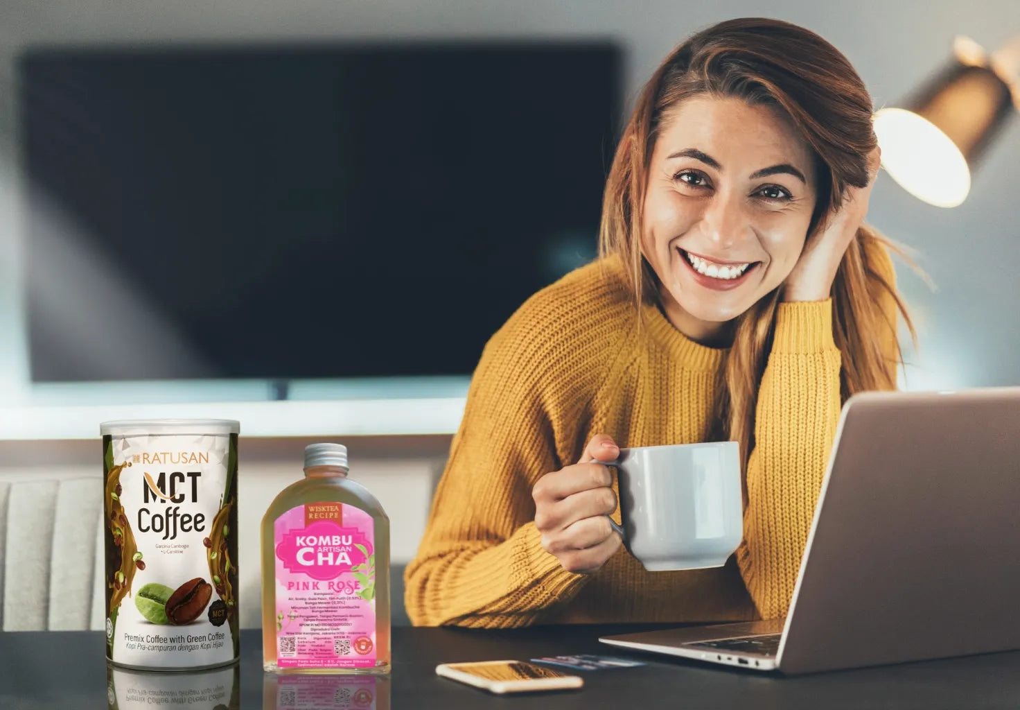 Woman in yellow sweater holding a mug with MCT Coffee and Kombu Cha products on a desk.